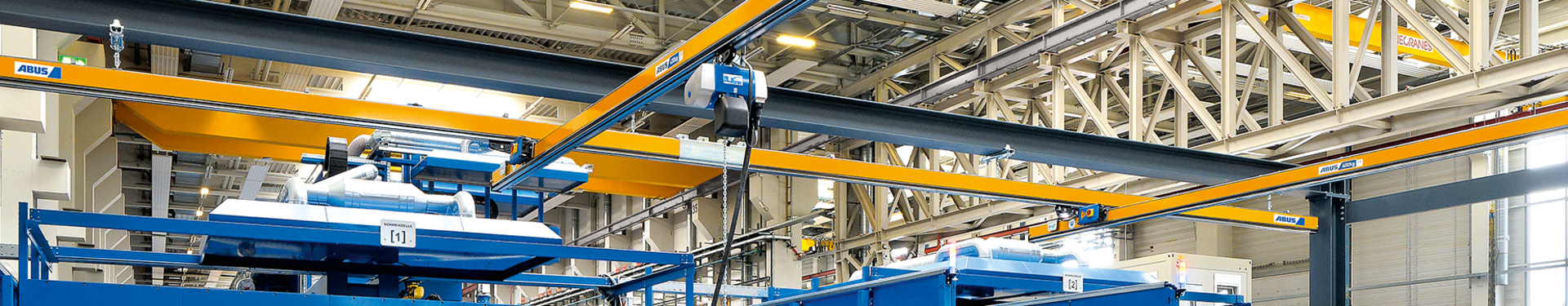 In un capannone industriale sono visibili diversi trasportatori aerei monotrave del tipo EHB. In primo piano un operaio che aziona un paranco a catena con un pulsante sospeso. Il paranco a catena scorre su un carrello a rulli nel profilo della trave principale.