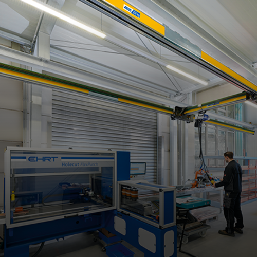 La gru monotrave rialzata EHB-X movimenta un carico all'interno di un capannone industriale, mentre sullo sfondo un operaio lavora a una macchina.